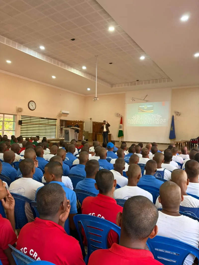 A large hall of CRPF recruits in training kit seated facing a Wealth Farming slide, addressed from the stage
